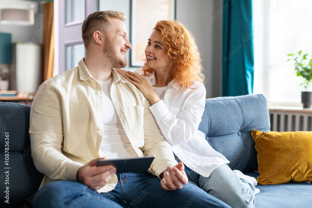 Cheerful loving spouses using digital tablet for shopping online, looking at each other and smiling, couple relaxing on couch in living room Cheerful loving spouses using digital tablet for shopping online, looking at each other and smiling, couple relaxing on couch in living room