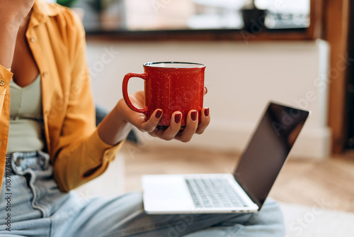 African American woman holding red cup of coffee working with laptop at home selective focus on hand