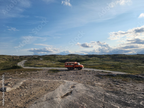 An orange vehicle is parked on a rocky terrain, surrounded by vast green fields and distant mountains. The sky is bright with clouds, creating a peaceful atmosphere. photo