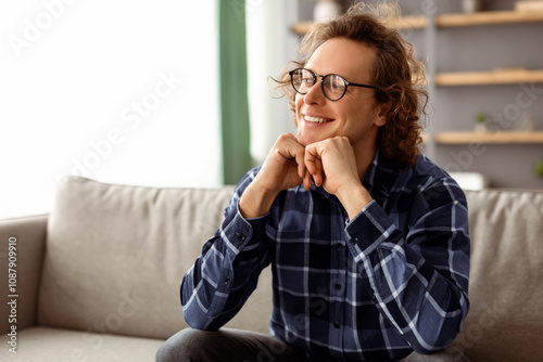 Positive Thinking. Happy Millennial Guy In Eyeglasses Sitting On Couch And Smiling Looking Aside At Home. Young Long-Haired Hipster Man Posing Indoors, Wearing Casual Clothes. Male Portrait Concept