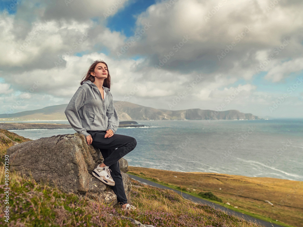 Teenager girl with long hair in grey hoodie is sitting on a rock overlooking stunning nature scenery of Achill island, Ireland.. The scene has a calm and peaceful mood. Travel and tourism concept. Teenager girl with long hair in grey hoodie is sitting on a rock overlooking stunning nature scenery of Achill island, Ireland.. The scene has a calm and peaceful mood. Travel and tourism concept.