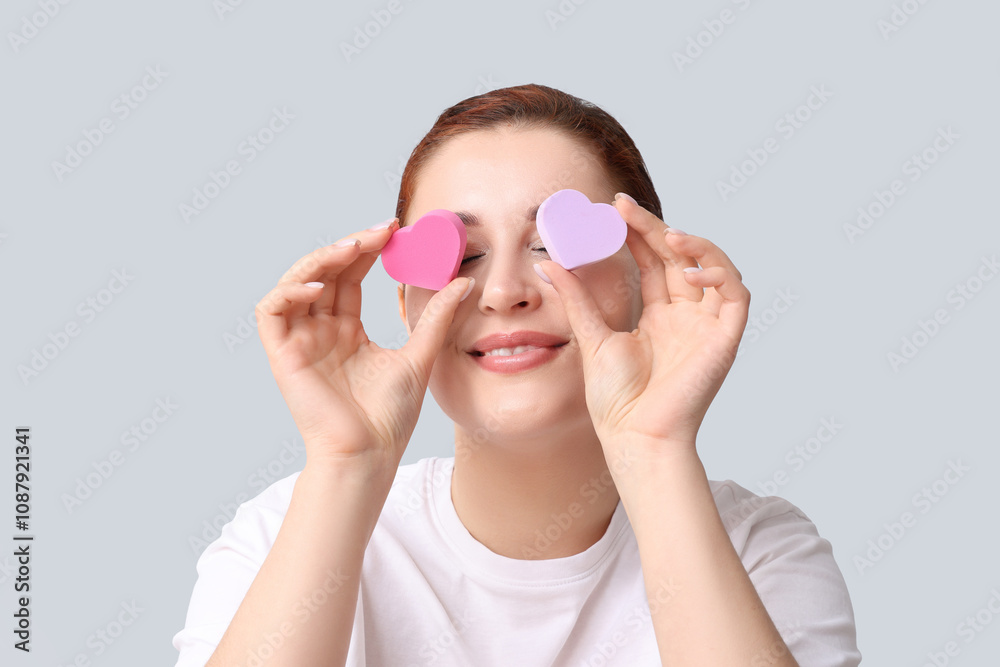 Beautiful young woman covering eyes with heart-shaped makeup sponges on white background Beautiful young woman covering eyes with heart-shaped makeup sponges on white background