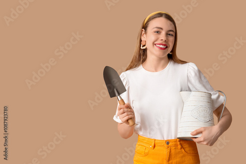 Beautiful young woman with watering can and gardening shovel on brown background