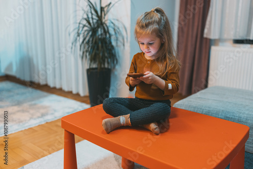 A young girl is sitting on an orange table, immersed in her smartphone, symbolizing technology’s role in modern childhood within a cozy home environment. photo