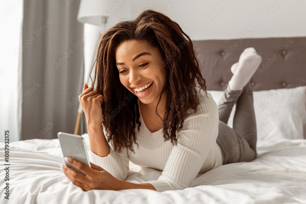 Big Win. Portrait Of Joyful African American Woman Holding Smartphone Shaking Fist In Joy And Excitement. Happy Young Female Lying In Bed At Home. Yeah, Great News And Message Concept