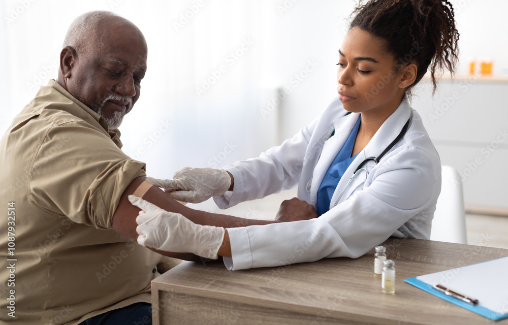 Fototapeta premium Vaccination Concept. Senior Black Man Getting Vaccinated Against Flu Or Corona Virus With Antiviral Vaccine, Female Doctor Putting Applying Sticking Adhesive Bandage On Arm After Injection In Hospital
