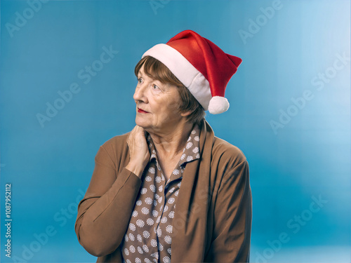 Obraz na płótnie Portrait of elderly beautiful woman with red Santa Claus hat isolated on blue background, festive people, Christmas template, copy space area w salonie