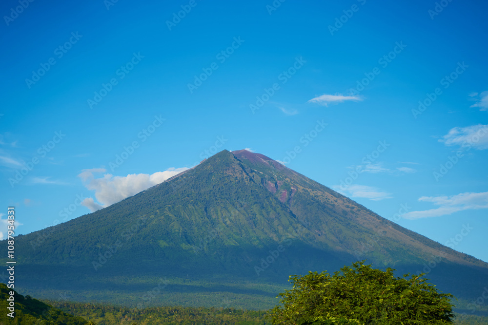 Panorama of Mount Agung and rice fields on the island of Bali. View of the mountain against a background of palm trees and a cornfield. Panorama of Agung volcano covered with clouds on a sunny day. Panorama of Mount Agung and rice fields on the island of Bali. View of the mountain against a background of palm trees and a cornfield. Panorama of Agung volcano covered with clouds on a sunny day.