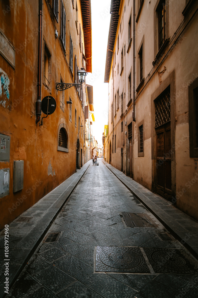 Two tourists walking down a narrow cobblestone street in florence, italy, with tall buildings on each side, towards a bright light at the end of the street Two tourists walking down a narrow cobblestone street in florence, italy, with tall buildings on each side, towards a bright light at the end of the street