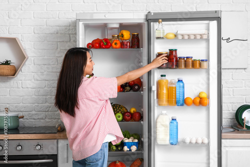 Young Asian woman taking jar of preserves from open fridge full of fresh food in kitchen photo