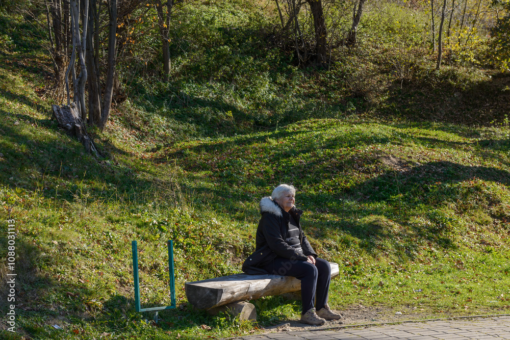 Elderly woman is sitting on log bench in Skhidnytsia, Ukraine. Elderly woman is sitting on log bench in Skhidnytsia, Ukraine.