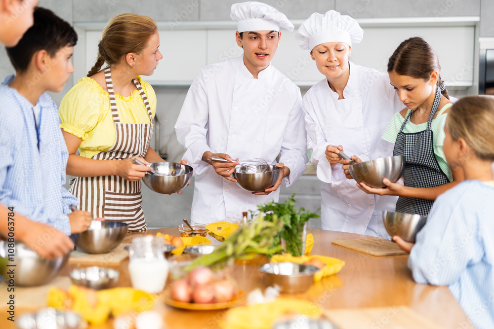At culinary master class, experienced male chef in white uniform explains to children subtleties of dessert preparation and female assistant helps child to work with whisk At culinary master class, experienced male chef in white uniform explains to children subtleties of dessert preparation and female assistant helps child to work with whisk