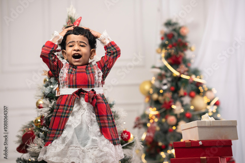 happy, smiling, pretty little girl wearing red check pattern Christmas dress touching her head over Christmas background with gift boxes, cheerful girl celebrating 2024 Merry Xmas and New Year.