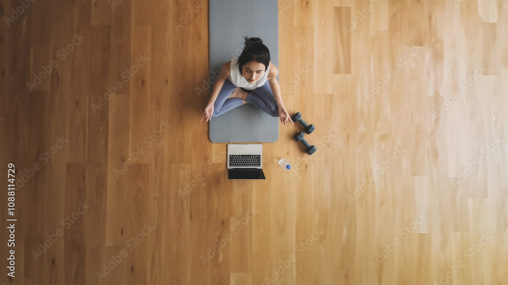 High angle view of young Asian woman practicing yoga and meditation at home sitting on floor in living room in lotus position with laptop and dumbbell. Mindful meditation and wellbeing concept High angle view of young Asian woman practicing yoga and meditation at home sitting on floor in living room in lotus position with laptop and dumbbell. Mindful meditation and wellbeing concept