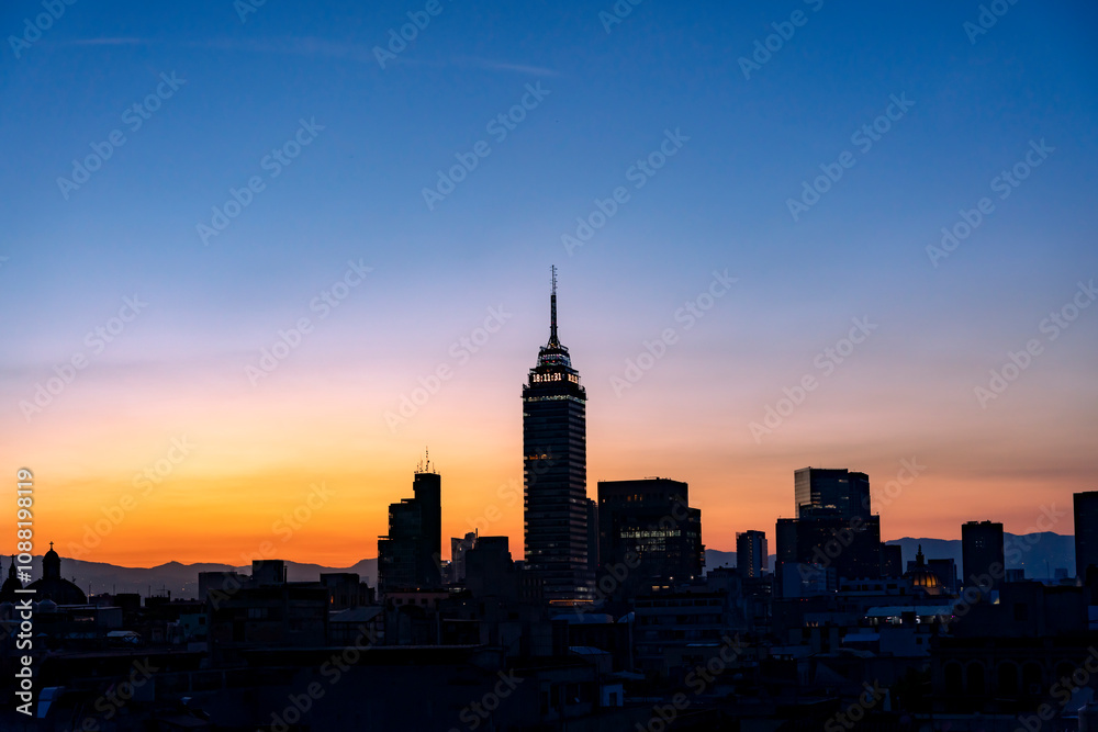 Obraz premium The Mexico City skyline at sunset, featuring Torre Latinoamericana and other buildings silhouetted against a colorful orange and blue sky.