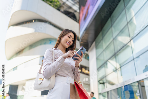 Asian Businesswoman Carrying Shopping Bags and Using Phone Amid Urban Cityscape Background