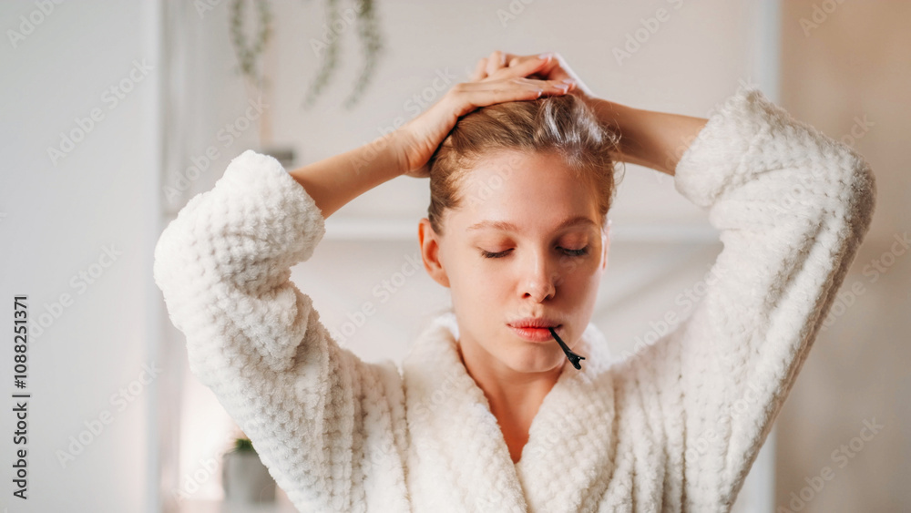 Beauty practice. Treatment procedure. Attractive young woman wearing white soft robe tying hair with hands with barrette in mouth. Beauty practice. Treatment procedure. Attractive young woman wearing white soft robe tying hair with hands with barrette in mouth.