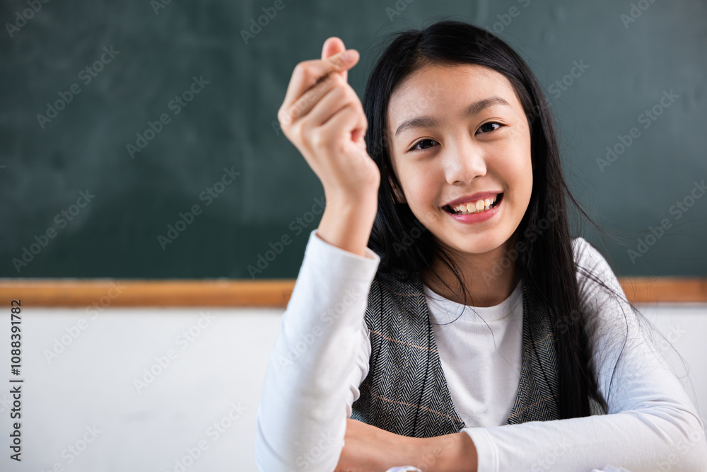 A young girl is smiling and holding her hand up in front of a blackboard. She is wearing a white shirt and a gray vest A young girl is smiling and holding her hand up in front of a blackboard. She is wearing a white shirt and a gray vest