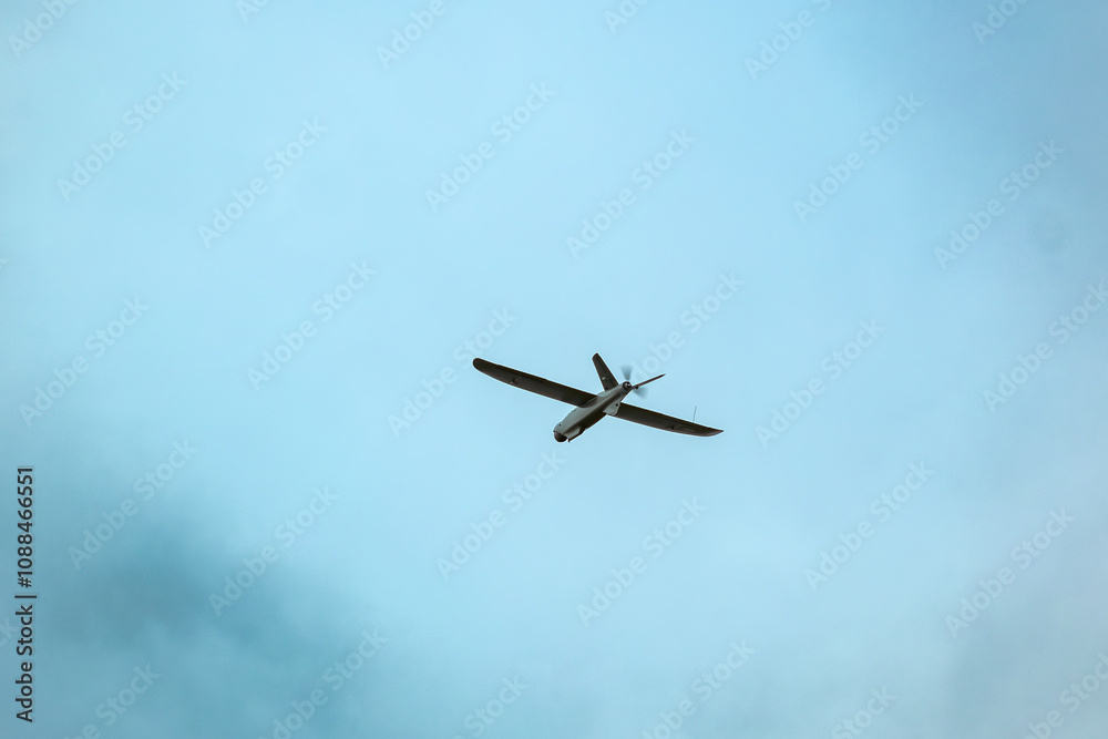 A drone flies against a bright blue sky with scattered clouds, surrounded by blurred green leaves in the foreground. aerial reconnaissance work A drone flies against a bright blue sky with scattered clouds, surrounded by blurred green leaves in the foreground. aerial reconnaissance work