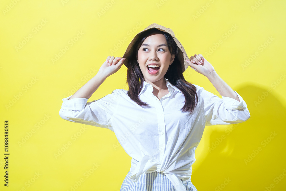 Asian woman wearing a white shirt and striped skirt, playfully holding a beige bucket hat with a cheerful expression against a vibrant yellow background, radiating joy and positivity. Asian woman wearing a white shirt and striped skirt, playfully holding a beige bucket hat with a cheerful expression against a vibrant yellow background, radiating joy and positivity.