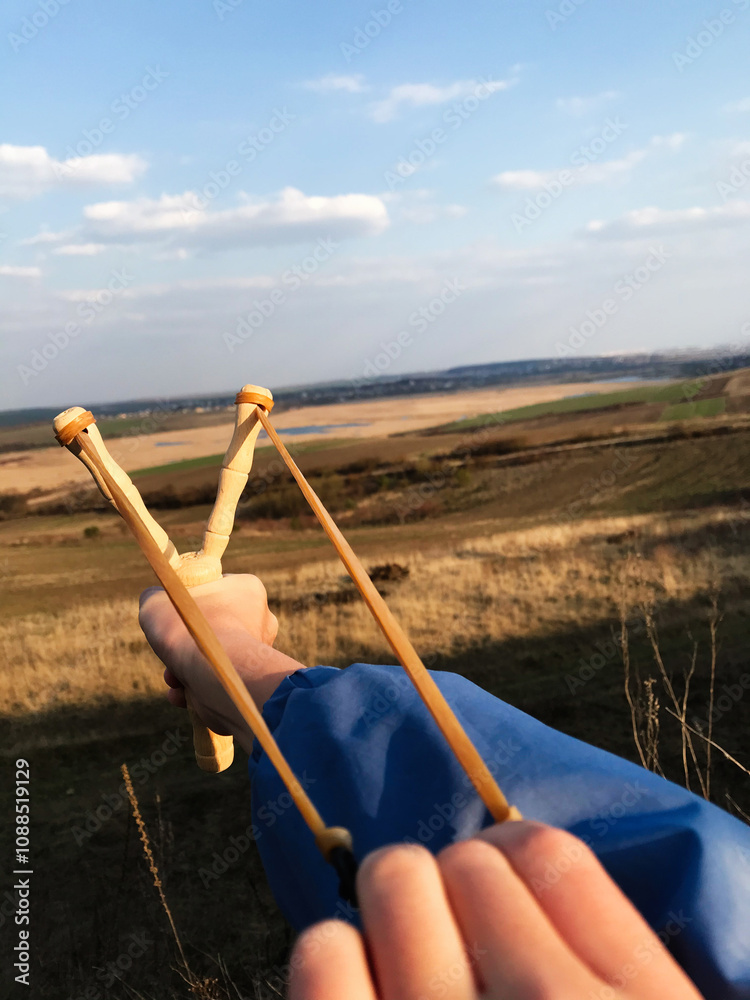 Fototapeta premium Close-up of hand aiming wooden slingshot in sunny landscape