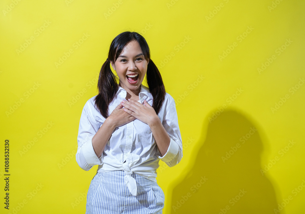 Asian woman smiling warmly, with one hand placed on her chest in a sincere gesture. She wears a white shirt and striped pants, posing against a bright yellow background