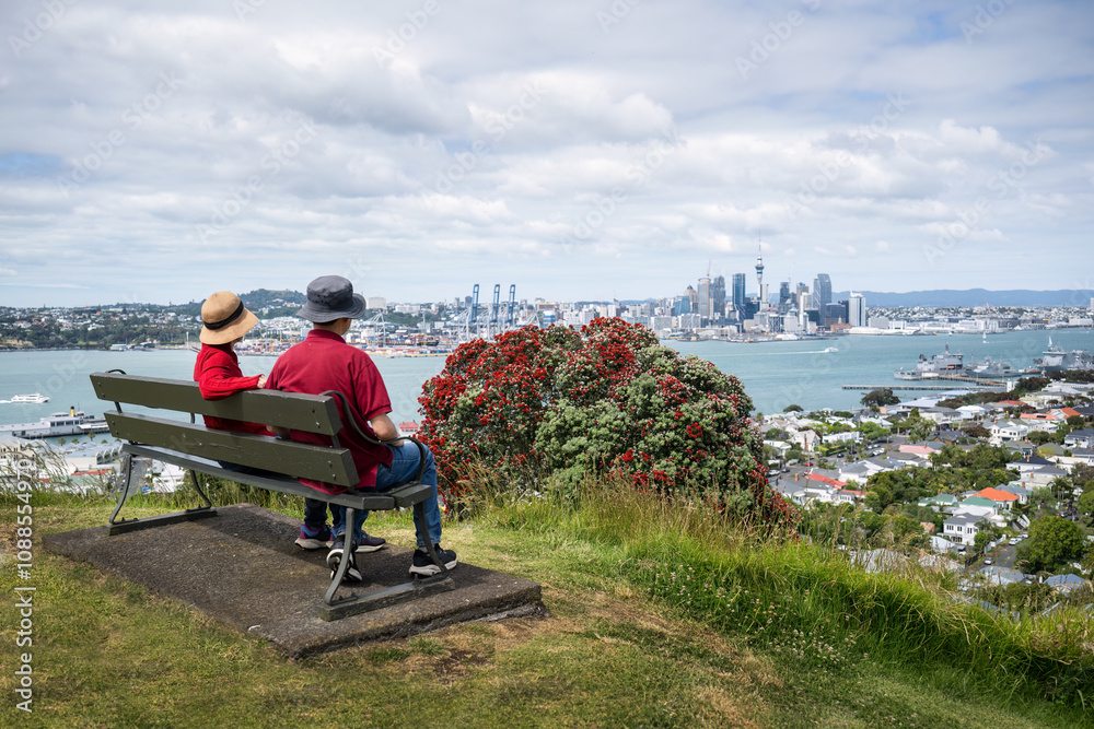 Couple sitting on the bench and enjoying the views of Auckland Sky Tower and skyline. Pohutukawa trees in full bloom.