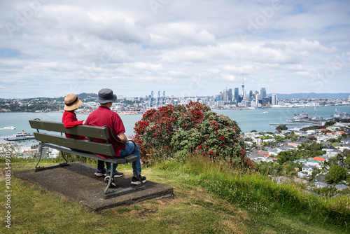 Obraz na płótnie Couple sitting on the bench and enjoying the views of Auckland Sky Tower and skyline. Pohutukawa trees in full bloom. w salonie