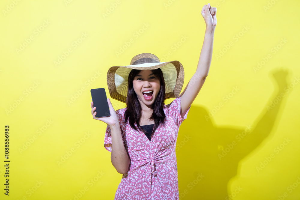 Asian woman in a pink floral dress, wearing a wide-brimmed straw hat, holding a smartphone in one hand and raising her other hand in celebration, smiling brightly against a vibrant yellow background.