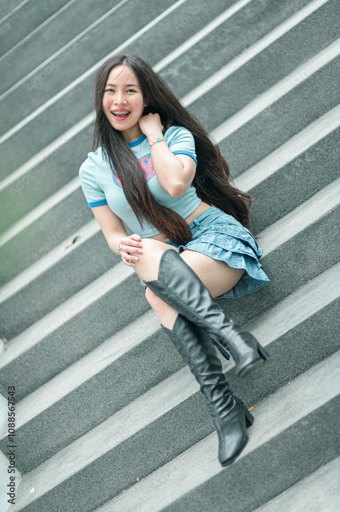 Obraz premium Young woman smiling while sitting on outdoor staircase, wearing stylish outfit with long hair and knee-high boots, showcasing confidence and modern fashion trends