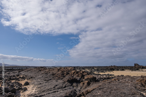 White sand beach with solidified volcanic lava flows, black rocks. Mountains in the background and sky with big white clouds. Caletón Blanco, Lanzarote, Canary Islands, Spain