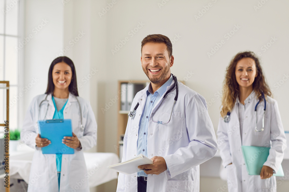 Fototapeta premium Medical team. Portrait of friendly Caucasian young male chief doctor with team in hospital office. Man in white medical coat together with two female colleagues practicing doctors smiling at camera.