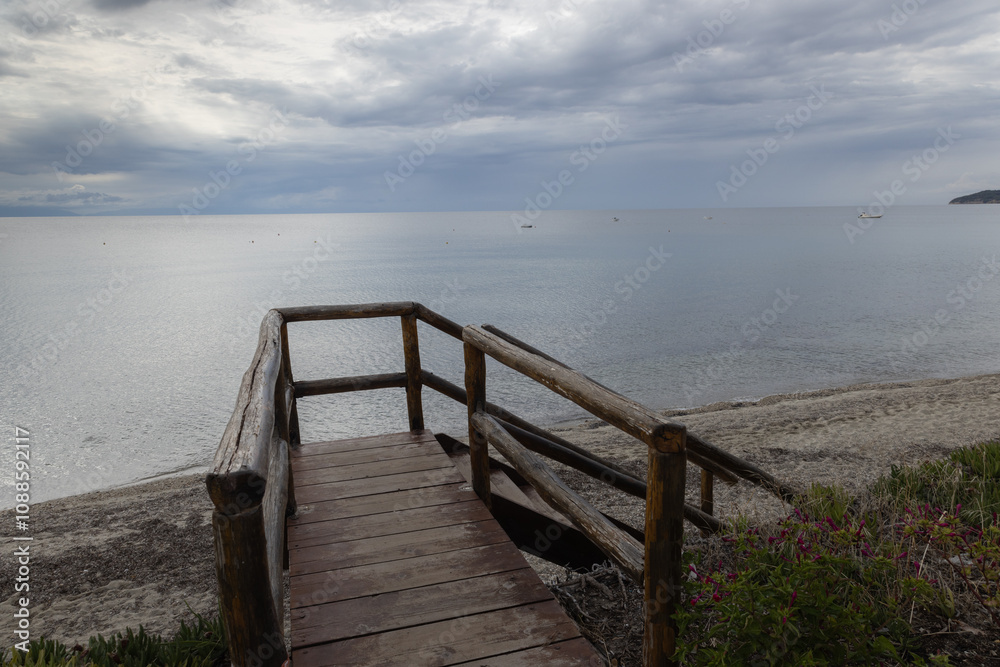 A serene beach scene with a wooden walkway leading to a calm sea under a cloudy sky, capturing the peaceful ambiance and natural beauty of the seaside setting. A serene beach scene with a wooden walkway leading to a calm sea under a cloudy sky, capturing the peaceful ambiance and natural beauty of the seaside setting.
