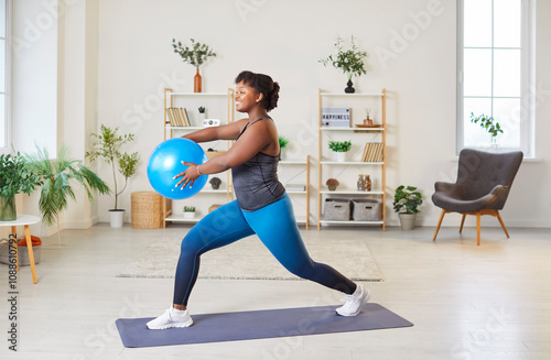 African American woman performing lunges with a fitball as part of her exercise routine at home gym. She focuses on fitness, training in a gymnastic style, and maintaining her workout regime. photo