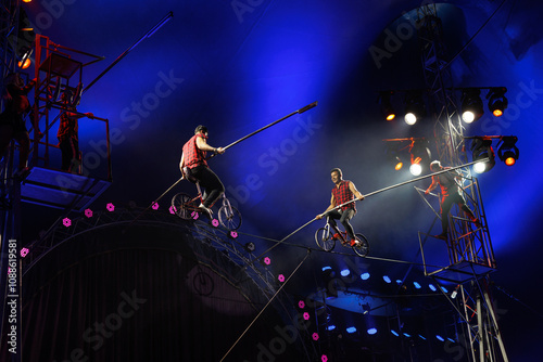 Two acrobats ride a bicycle on a tightrope under the dome of the circus.