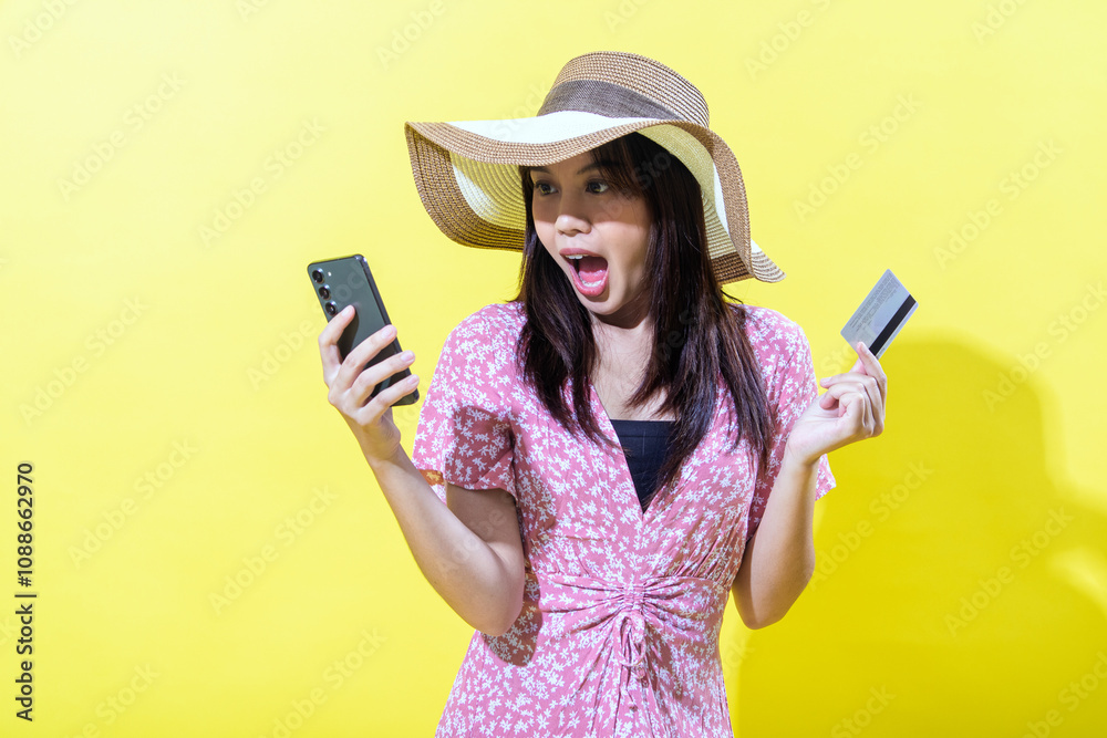 Asian woman in a pink floral dress, wearing a wide-brimmed straw hat, holding a smartphone and a credit card with a surprised expression against a vibrant yellow background, showcasing shock Asian woman in a pink floral dress, wearing a wide-brimmed straw hat, holding a smartphone and a credit card with a surprised expression against a vibrant yellow background, showcasing shock