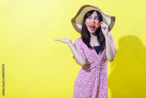 Young Asian woman in a floral pink dress posing cheerfully with a wide-brimmed straw hat and pink sunglasses, standing against a bright yellow background, expressing excitement and joy