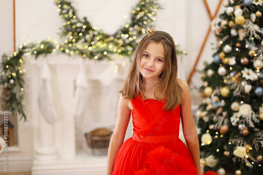 Smiling Girl in Red Dress by Christmas Tree. A young girl wearing a red dress and a golden crown smiling warmly near a beautifully decorated Christmas tree and garland. Smiling Girl in Red Dress by Christmas Tree. A young girl wearing a red dress and a golden crown smiling warmly near a beautifully decorated Christmas tree and garland.