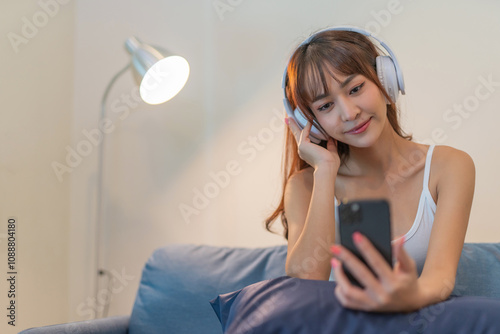 Beautiful woman wearing headphone listening to music on her smartphone, sitting on sofa in her room relaxingly photo