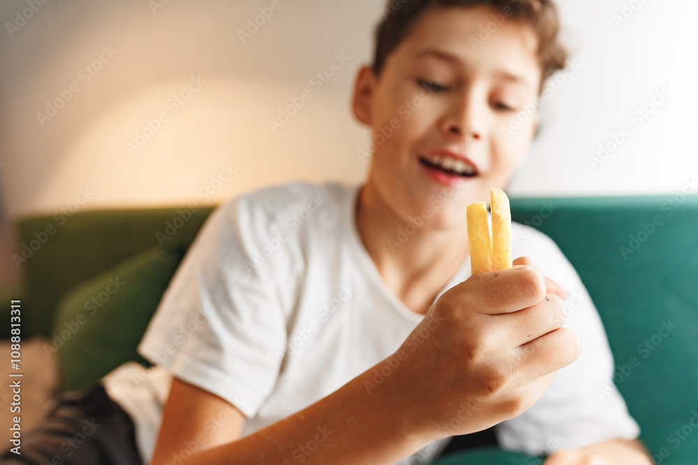 Close up portrait of a satisfied cute boy eating french fries at home on sofa while reading or doing homework. Unhealthy food concept, close up Close up portrait of a satisfied cute boy eating french fries at home on sofa while reading or doing homework. Unhealthy food concept, close up