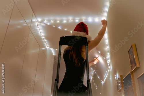 A woman in a Santa hat stands on a ladder, hanging and instaling Christmas lights on the wall of her home. She carefully arranges the festive lights, creating a warm holiday atmosphere. Stock photo photo