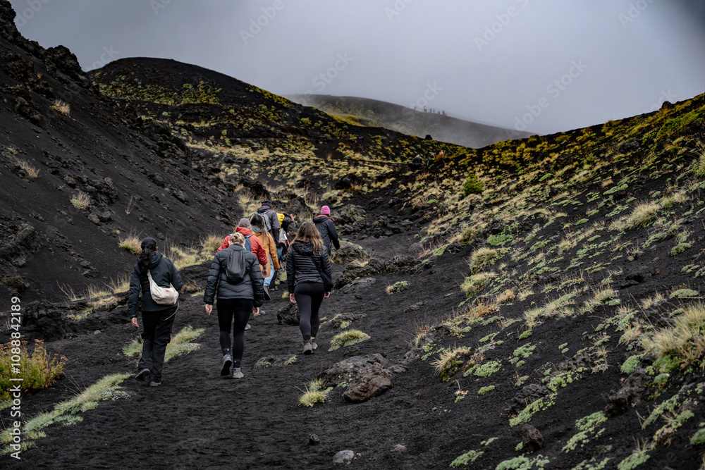 Obraz premium Photography of A group of hikers in outdoor gear hiking and climbing the dark and rocky, barren slopes of Mount Etna, Sicily. during a foggy and cloudy day. Stock photo