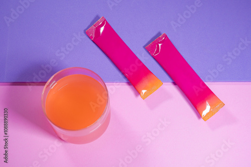 A glass of orange water placed on a pastel purple and pink background, and colored packets, primarily pink with a gradient that transitions to orange