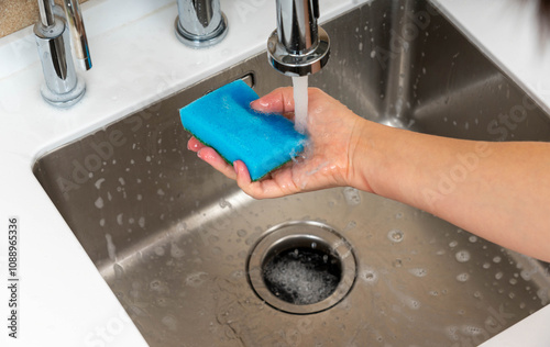 A close-up of a blue sponge being washed under running water in a stainless steel kitchen sink, ensuring cleanliness and hygiene.