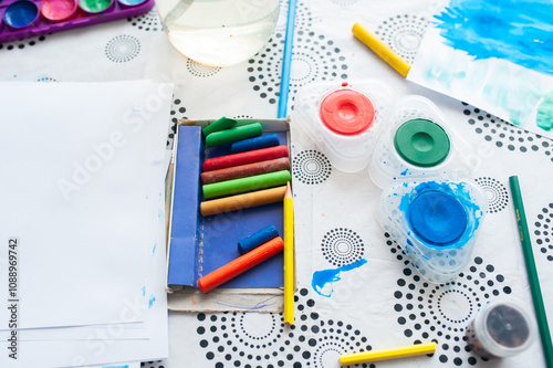 A set of drawing accessories. On the table is a white sheet of paper, watercolor paints, pencils