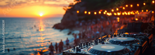 A close-up shot of hands on turntables, with people dancing in soft focus behind at sunset during an outdoor party. A photo of DJ equipment on the beach with a sunset and crowd in the background.