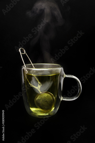 Double transparent glass mug, with green tea and silver clamp tea strainer, visible steam, isolated on black background