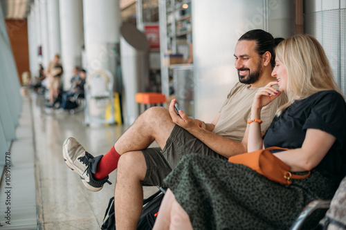 A man and a woman are sitting at the boarding gate at the airport. A married couple is waiting to board a plane at a large window near the gate photo