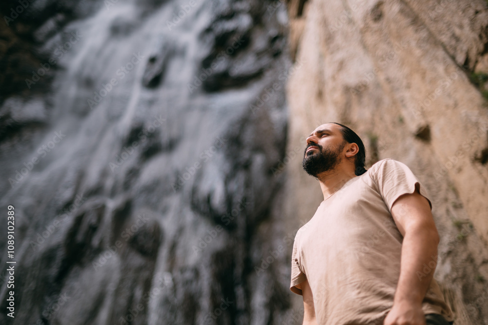 Fototapeta premium Portrait of a male tourist standing at a mountain waterfall. A young guy on the background of a large waterfall in the mountains