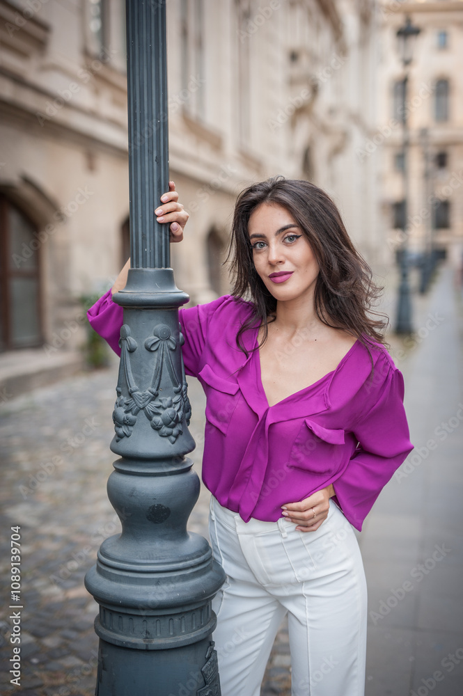 Beautiful young brunette woman in a purple blouse and white pants posing outdoor leans against a pillar .Portrait of a beautiful young brunette woman in a purple jacket posing